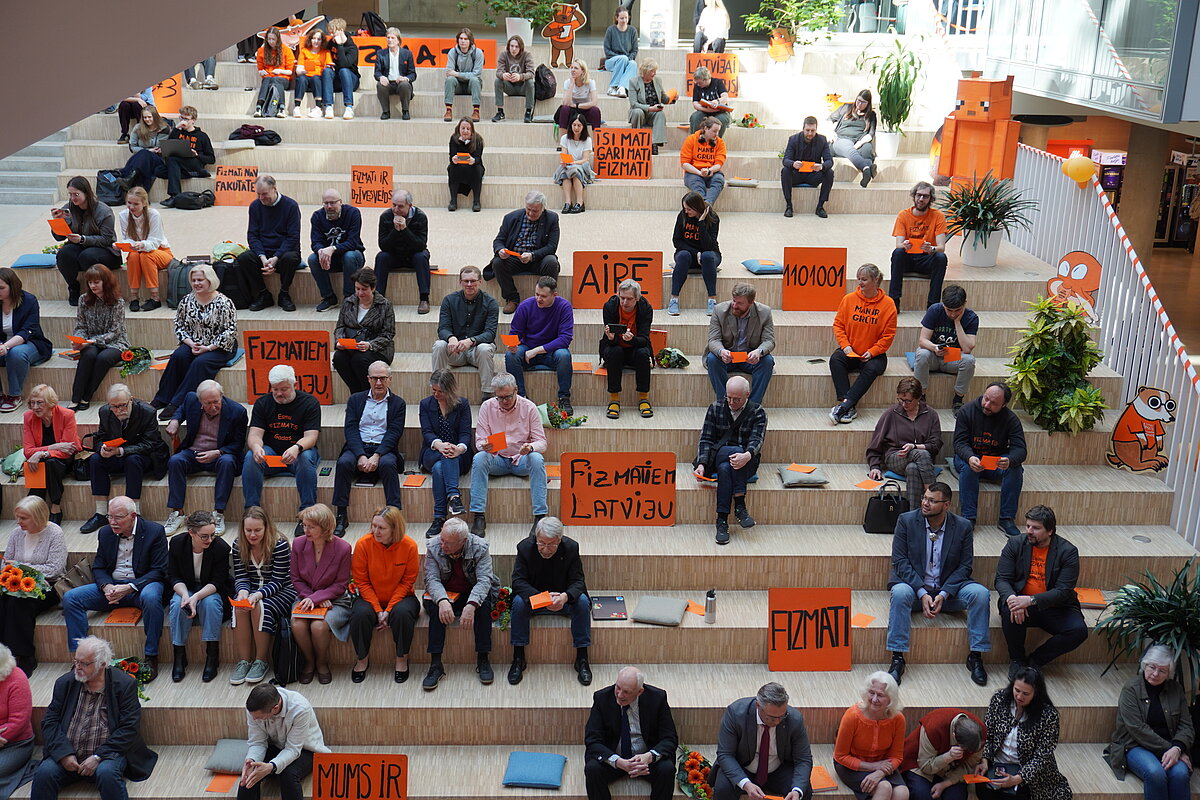 Plaša kadra fotogrāfija, kurā daudzi cilvēki sēž Zinātņu mājā uz koka amfiteātra tipa pakāpēm un rokās tur oranžus plakātus ar uzrakstiem latviešu valodā, tostarp “FIZMATI”, “Fizmatiem Latviju” un “AIPF”; telpā redzamas oranžas dekorācijas un augi.