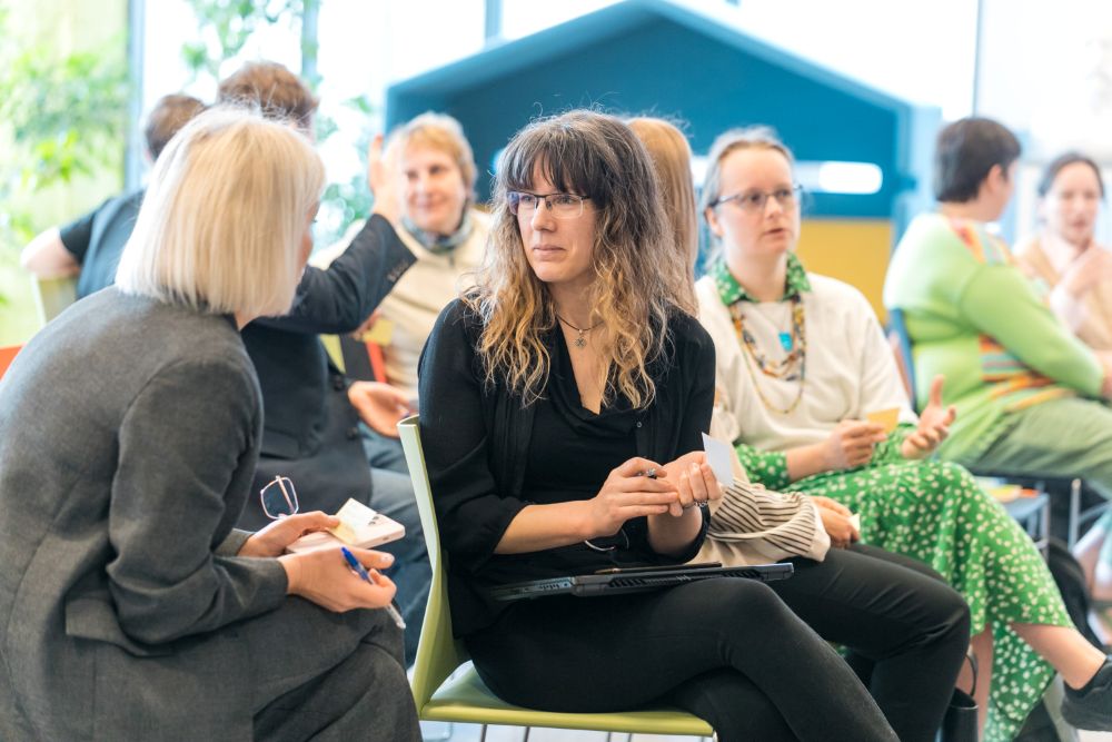 Cilvēku grupa sēž uz krēsliem telpā un iesaistās sarunā vai diskusijā. Priekšplānā divi dalībnieki sarunājas, turot piezīmes un pildspalvu, fonā redzami citi klausītāji