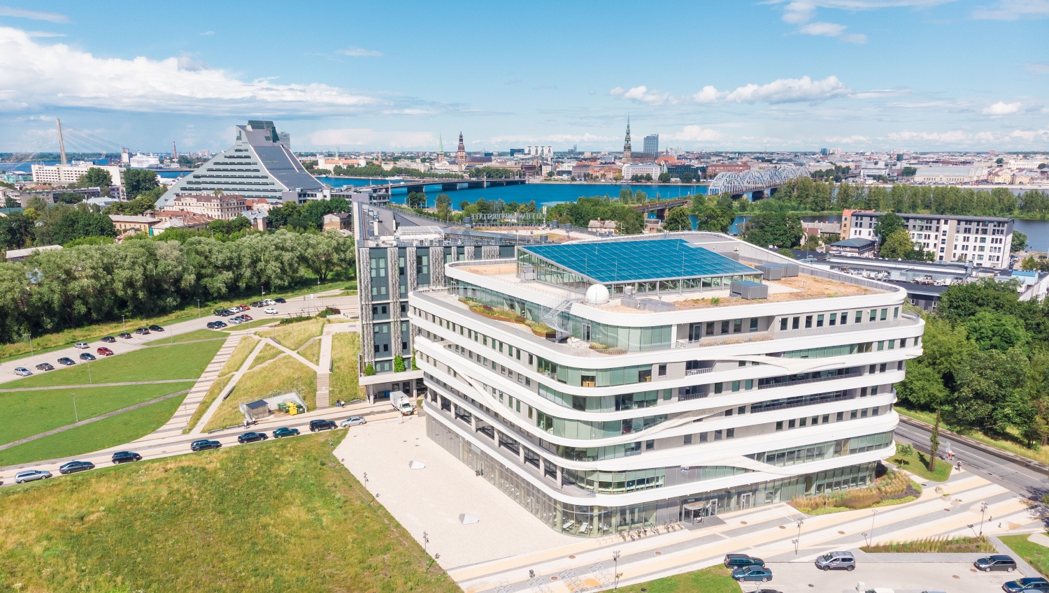 Skats no augšas uz mūsdienīgu daudzstāvu ēku ar stiklotām fasādēm un noapaļotām līnijām, kas atrodas zaļā teritorijā pie Daugavas; fonā redzams Rīgas pilsētas panorāmas siluets, tilti un ūdens, priekšplānā — ceļi, gājēju celiņi un apstādījumi.