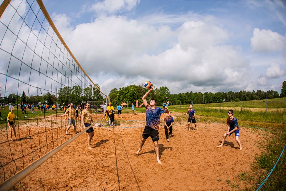 Pludmales volejbola spēle smilšu laukumā, kur vairāki spēlētāji piedalās izspēlē pie tīkla; viens spēlētājs paceļ bumbu sitienam. Fonā redzami skatītāji, zaļa ainava un mākoņainas debesis.