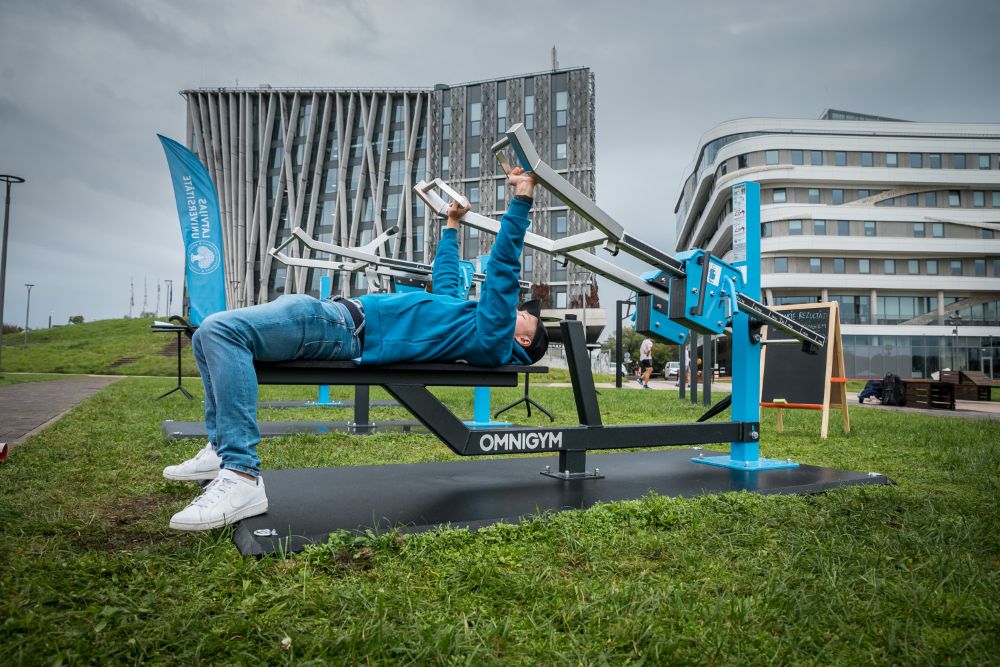 Cilvēks guļ uz āra trenažiera soliņa un paceļ svara stieni, sportojot brīvā dabā uz zāliena. Fonā redzamas LU Dabas māja un LU Zinātņu māja  un āra sporta iekārtas