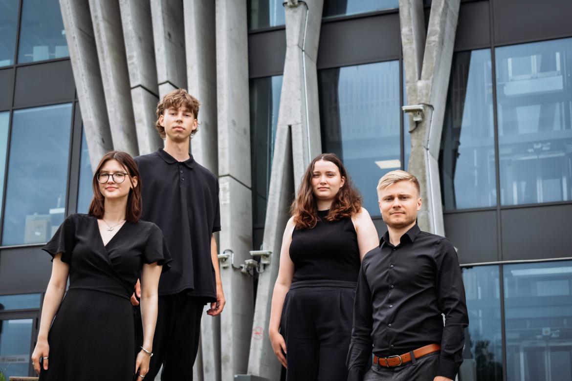 Četru personu grupa stāv brīvā dabā pie mūsdienīgas ēkas ar vertikāliem betona balstiem un stikla fasādi; visi ģērbušies tumšas krāsas apģērbā.