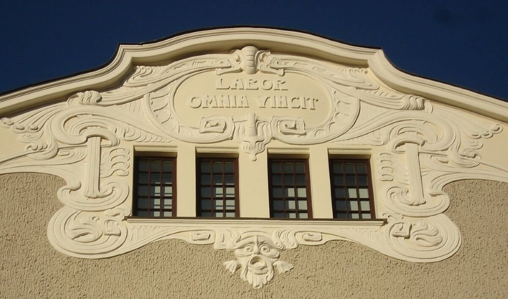 Ēkas fasādes augšdaļa ar bagātīgi veidotu dekoratīvu apmetuma reljefu, trīs logiem un centrā izvietotu latīņu uzrakstu “Labor omnia vincit”