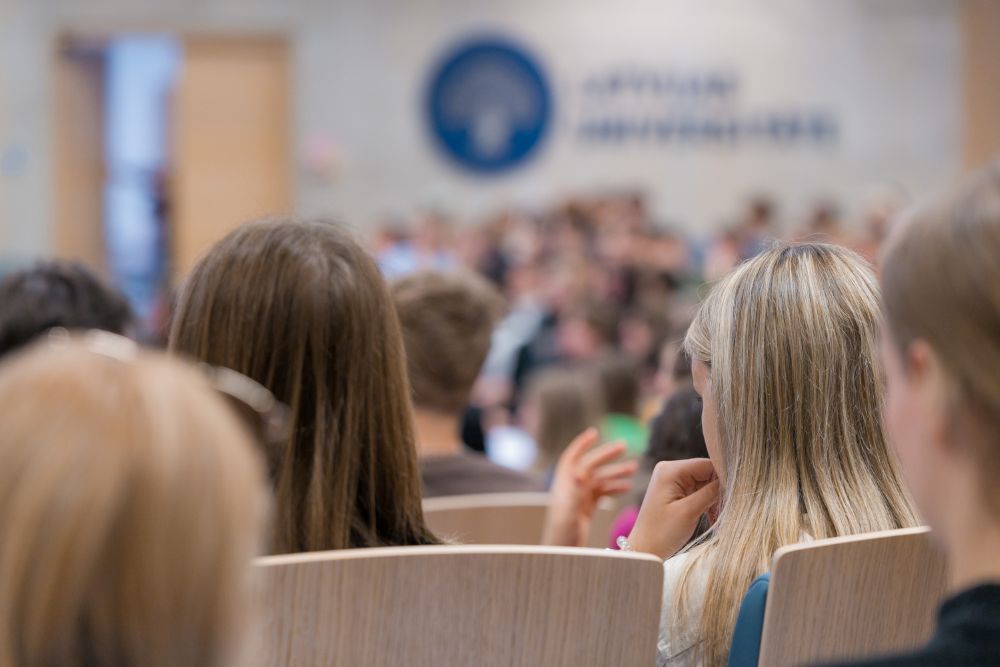 Cilvēki auditorijā, fonā LU logo