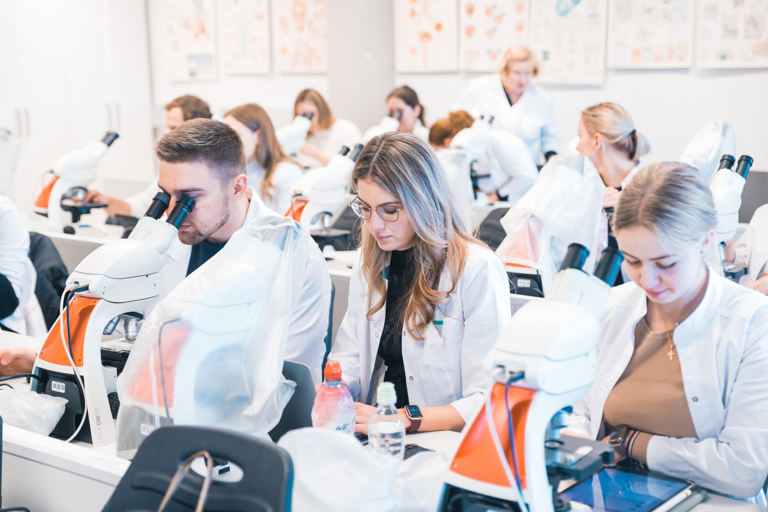 Studenti laboratorijā strādā ar mikroskopiem; visi tērpti baltos halātos un sēž pie darba galdiem ar laboratorijas aprīkojumu, fonā redzama mācību telpa ar plakātiem un pasniedzēju.