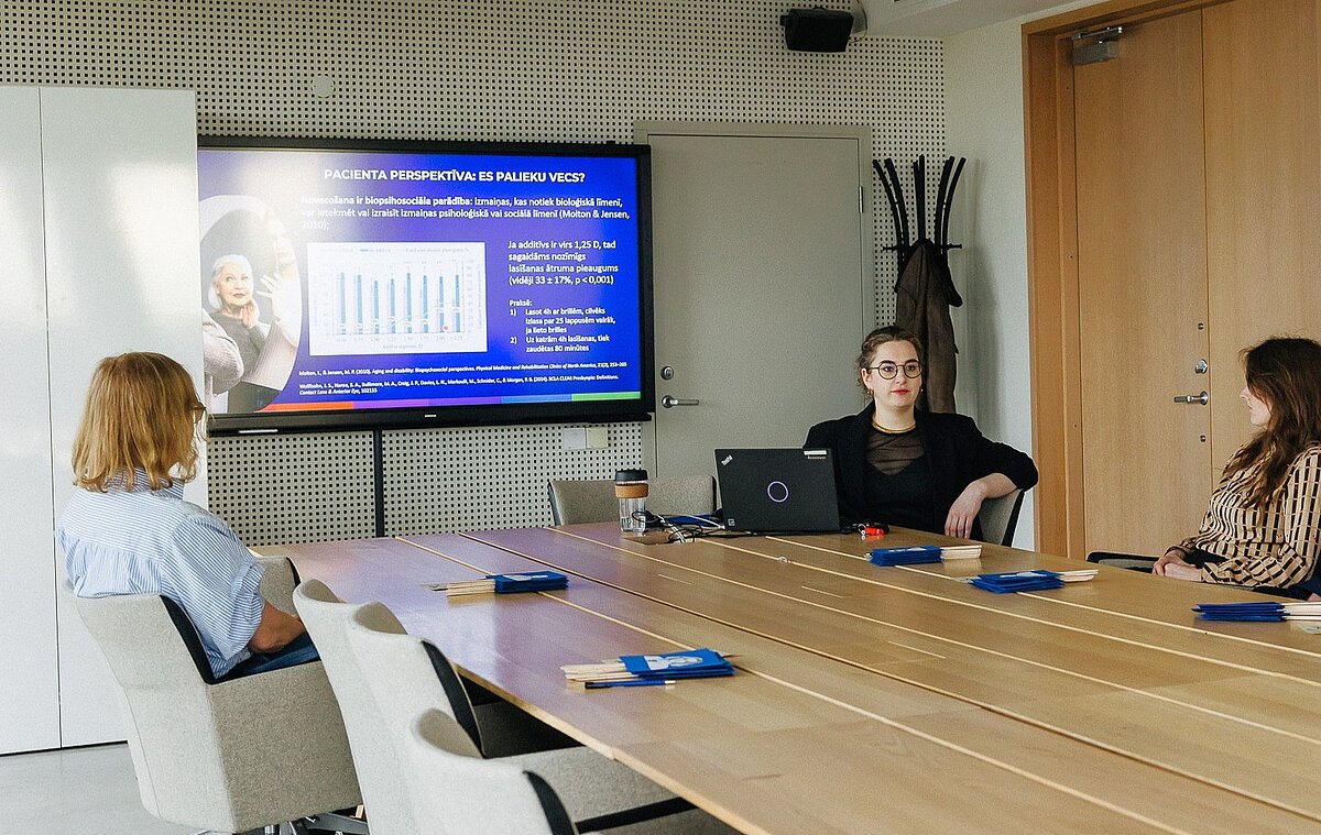 Neliela sanāksme vai prezentācija konferenču telpā; pie galda sēž vairākas personas, priekšā redzams ekrāns ar slaidu par pacientu perspektīvu un pētījuma datiem, centrā atvērts portatīvais dators, uz galda izvietoti informatīvi materiāli.
