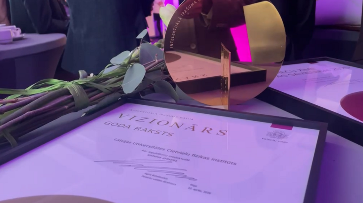 Apbalvošanas pasākuma galds ar pušķi, stiklotu balvas statueti un diplomu rāmī; priekšplānā redzams diploms ar uzrakstu “VIZIONĀRS – gada raksts”, fonā violets pasākuma apgaismojums un citi apbalvojumi.