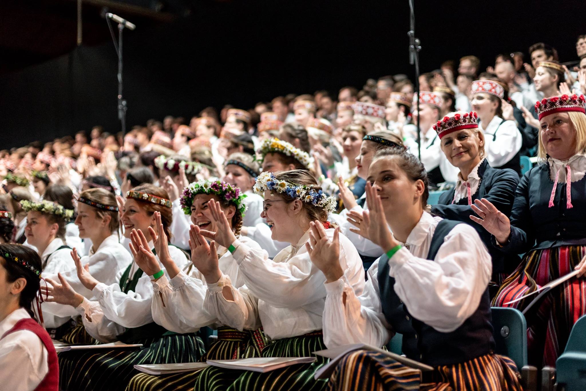 Cilvēku koris tautastērpos aplaudē koncertzālē.