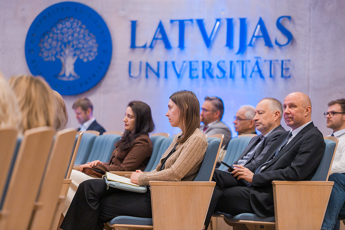 Cilvēki konferenču zālē, fonā Latvijas Universitātes logo