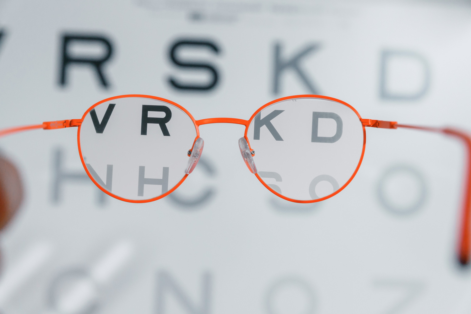 Spilgti oranžas brilles novietotas acu pārbaudes tabulas priekšā; caur briļļu lēcām burti redzami asi un skaidri (piemēram, “V”, “R”, “K”, “D”), bet ārpus lēcām tie ir izplūduši, ilustrējot redzes korekcijas efektu.