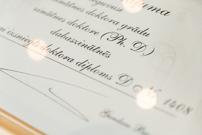 Doktora diploma tuvplāns ar tekstu “Ph. D.” un redzamu paraksta vietu.