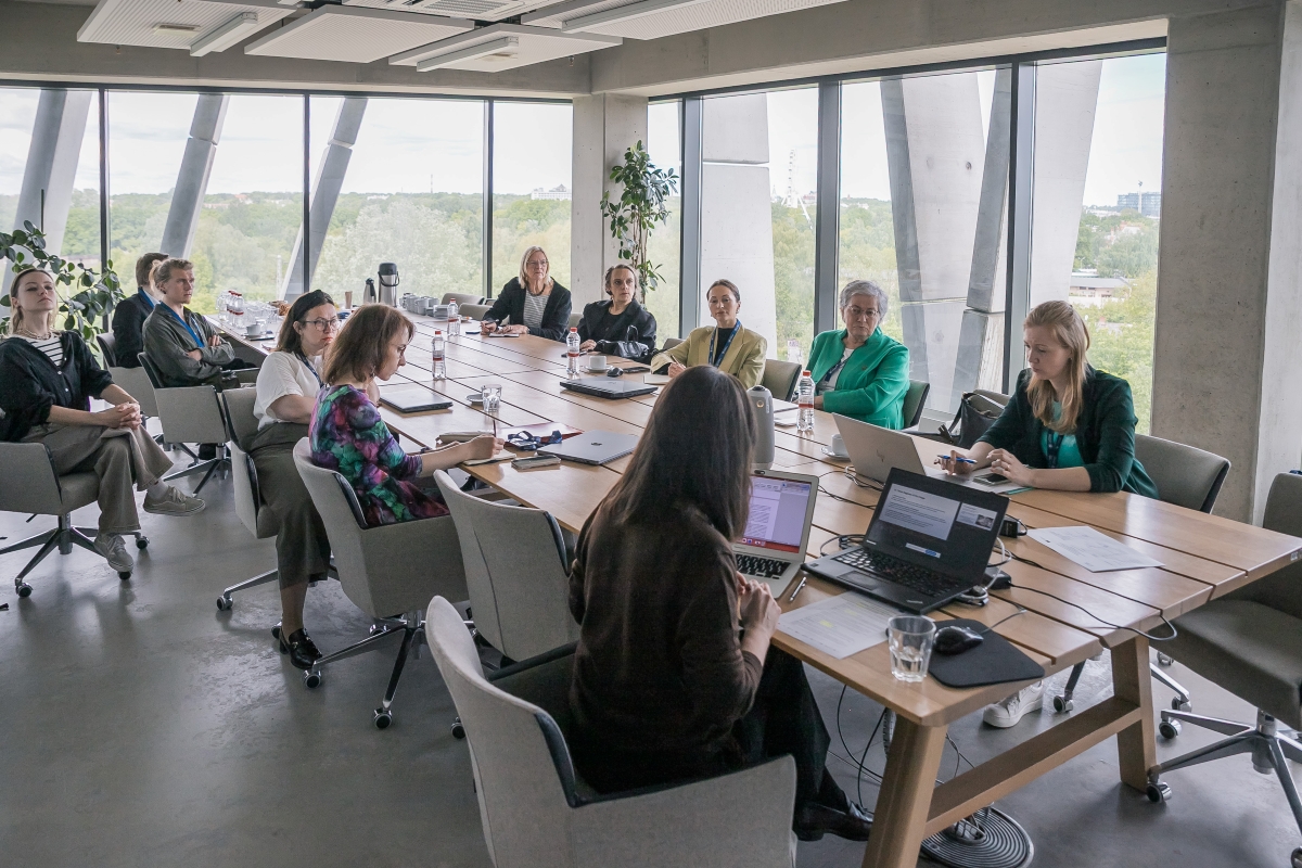 Cilvēku grupa sēž pie galda modernā sanāksmju telpā ar lieliem logiem un strādā pie portatīvajiem datoriem.