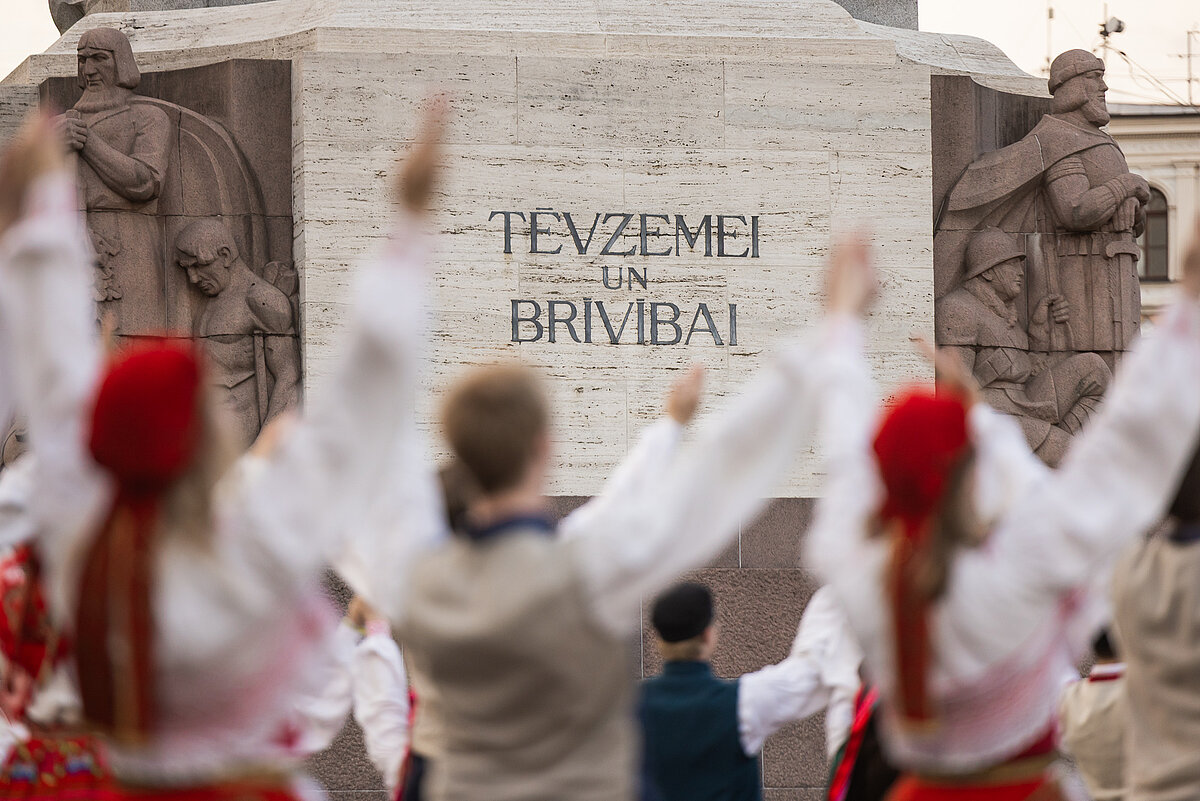 Tautastērpos ģērbti svētku dalībnieki pie Brīvības pieminekļa, fonā redzams uzraksts “Tēvzemei un Brīvībai”.