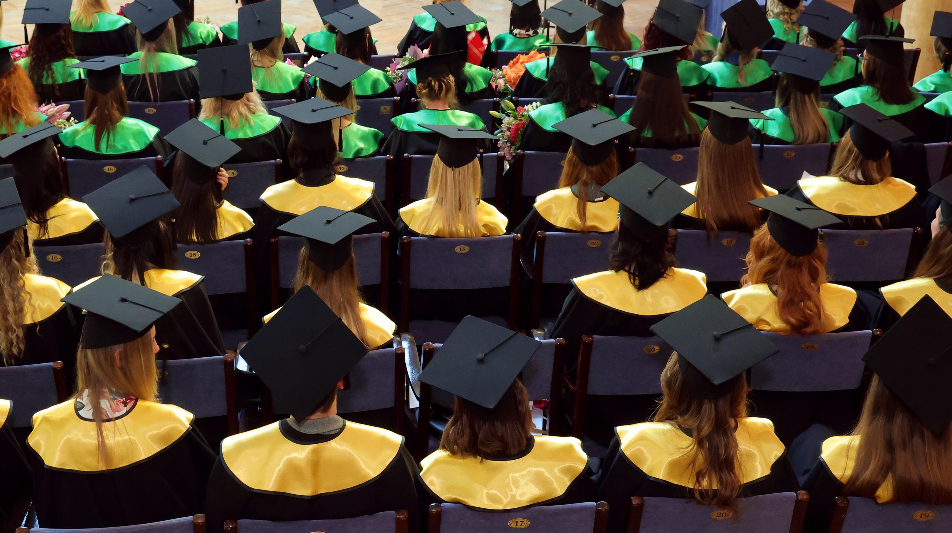 **Alt teksts:**   Auditorijā rindās sēž absolventi melnās cepurēs un talāros ar zaļām un zelta kakla lentēm, skatoties uz priekšu izlaiduma ceremonijas laikā. Dažiem rokās ziedi, redzamas krēslu rindas ar numuriem.
