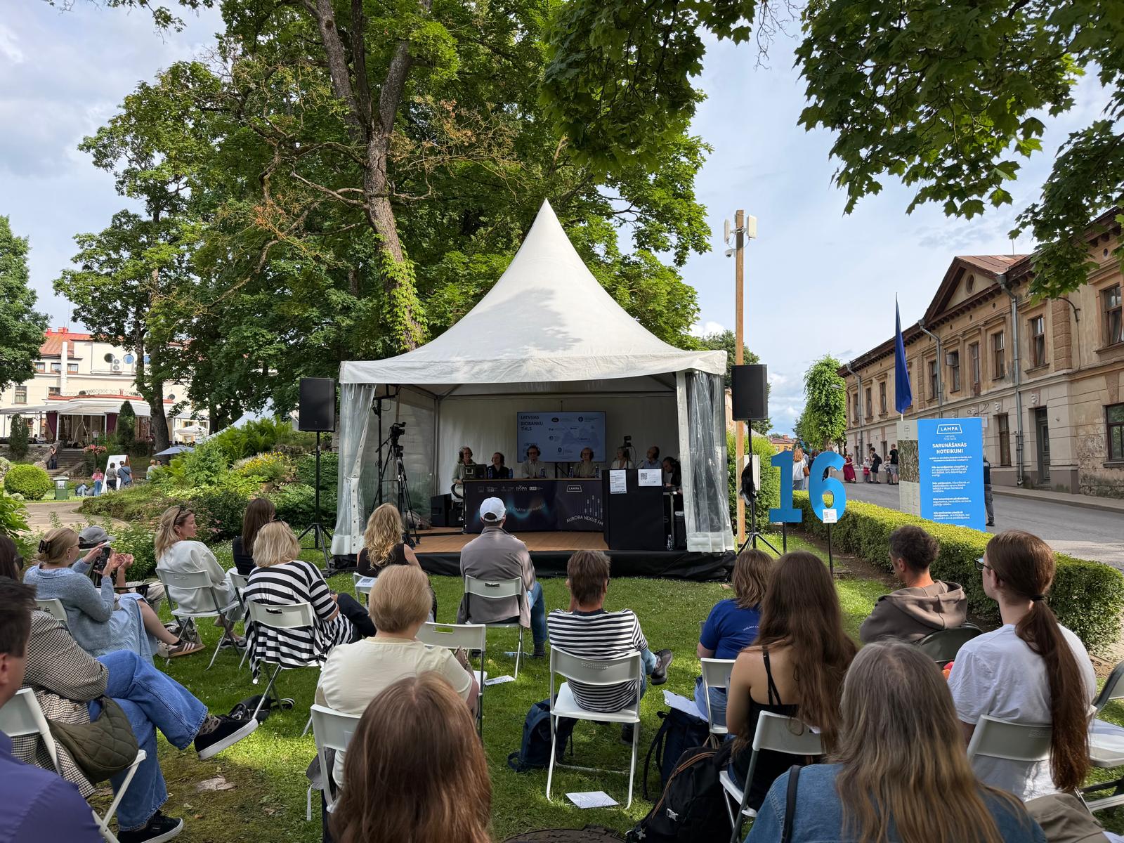 Brīvdabas diskusija festivālā “LAMPA”, kur klausītāji sēž krēslos zem kokiem un vēro sarunu uz skatuves zem baltas nojumes. Pasākums notiek pilsētvidē, apkārt redzami koki, ēkas un informatīvas norādes.
