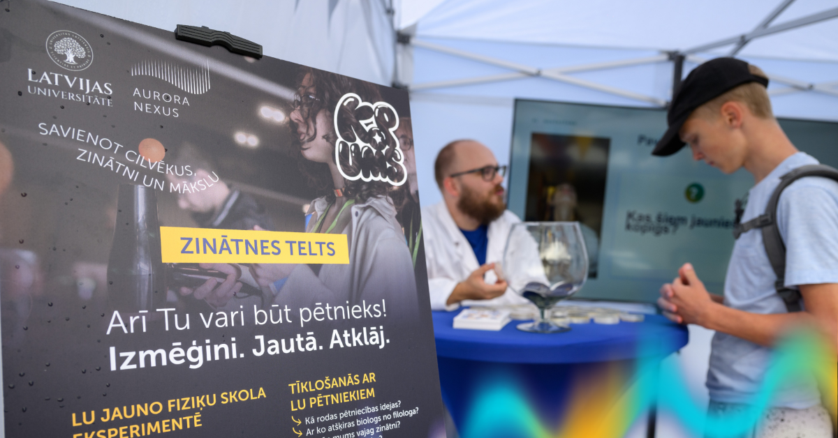 Informatīvs plakāts par Latvijas Universitātes un Aurora Nexus Zinātnes telti, bet fonā pie galda notiek saruna starp pasākuma apmeklētāju un zinātnes aktivitātes vadītāju.