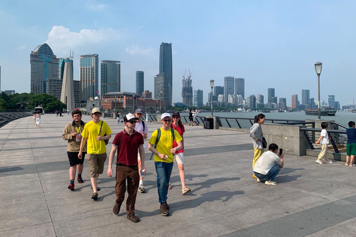 Skolēnu grupa pastaigājas pa promenādi pie upes, fonā redzami debesskrāpji un mūsdienīga pilsētas panorāma.
