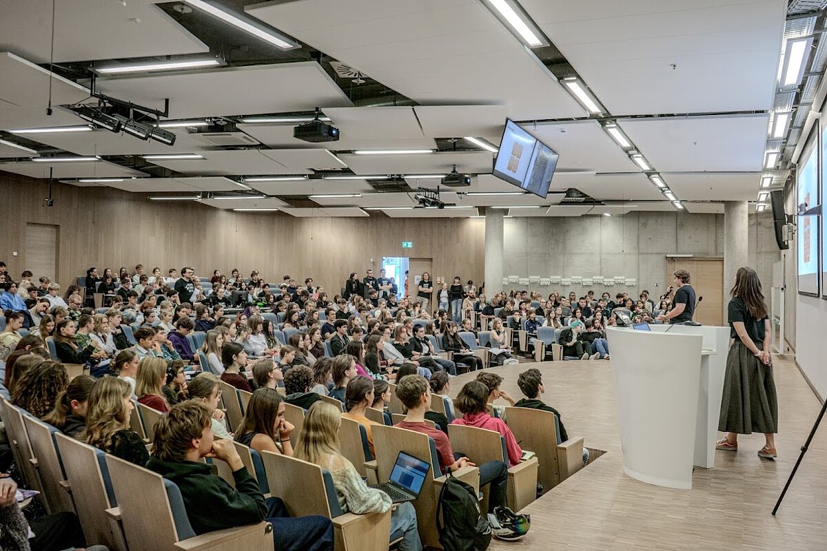 Alt teksts:  Pilna auditorija ar skolēniem un studentiem klausās prezentāciju modernā universitātes lekciju zālē.