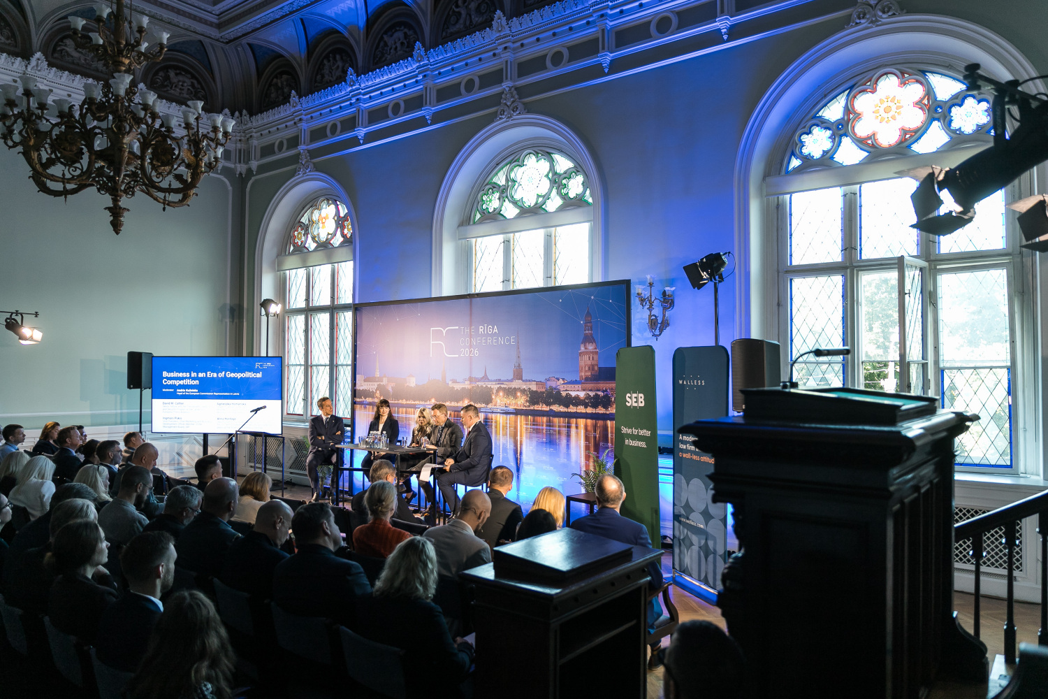 Alt teksts:  Konferences paneļdiskusija vēsturiskā zālē ar klausītāju auditoriju, skatuvi, prezentācijas ekrānu un izgaismotu fonu.