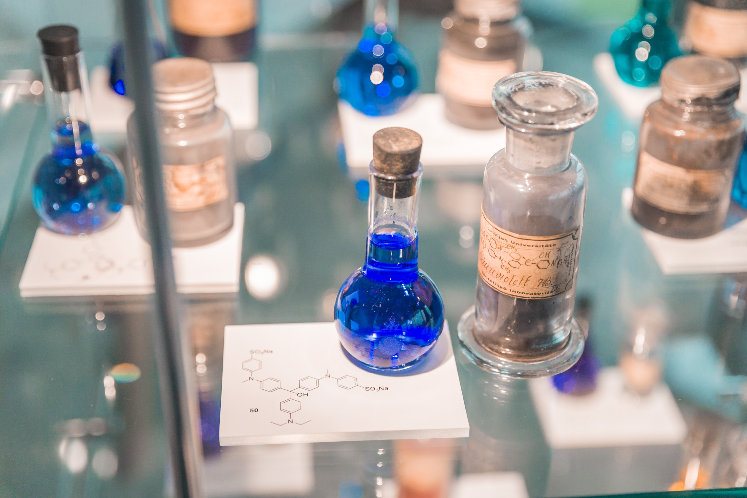 Stikla vitrīnā izstādīti dažādi laboratorijas stikla trauki ar šķīdumiem, tostarp kolbas un pudelītes ar ziliem un caurspīdīgiem šķidrumiem; priekšplānā neliela kolba ar zilu šķīdumu uz balta pamatnes ar ķīmiskās struktūras attēlu, fonā redzami vairāki vēsturiski marķēti reaģentu trauki.