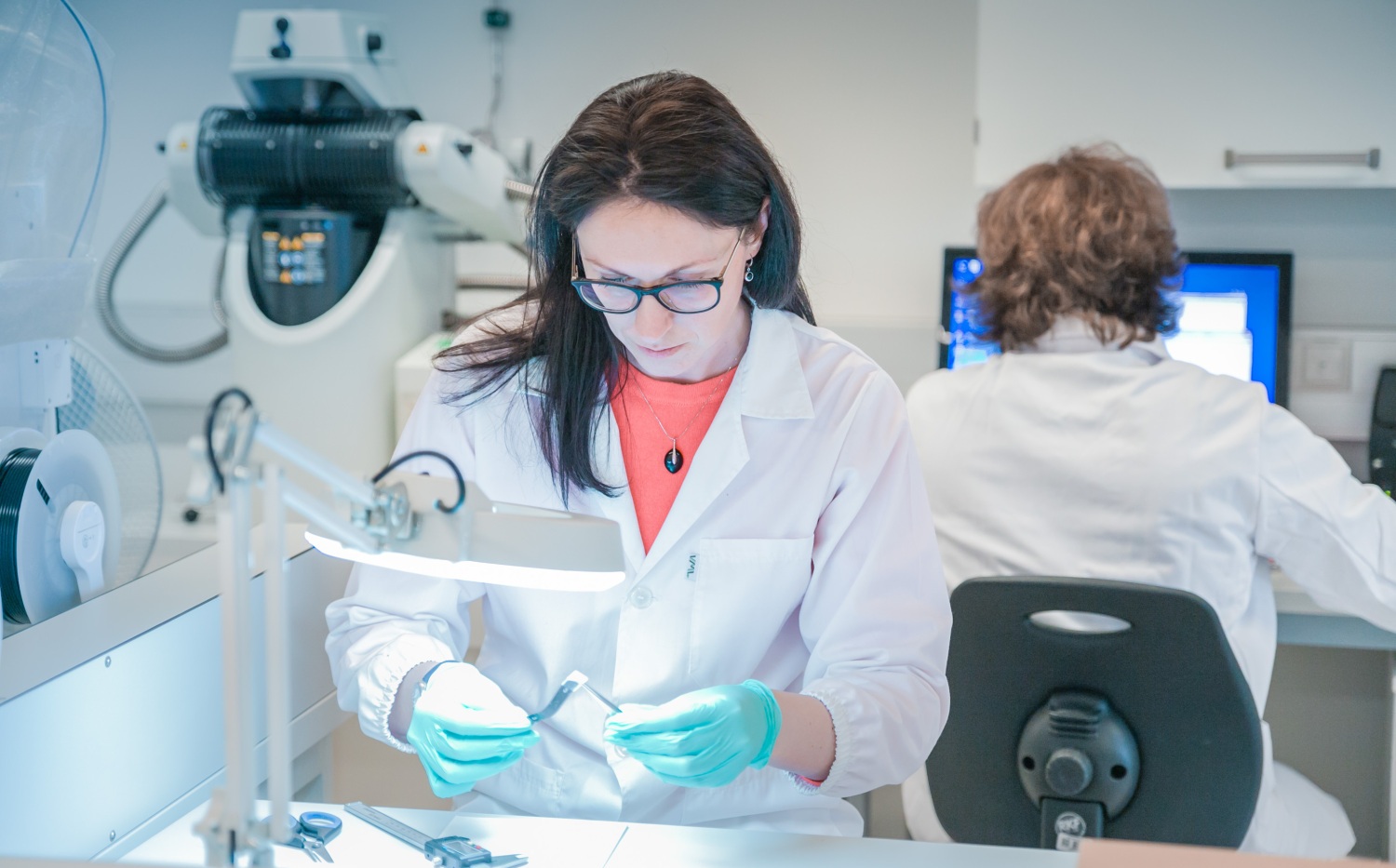 Persona laboratorijā baltā halātā un cimdos strādā ar nelielu paraugu pie darba galda; fonā redzama laboratorijas iekārta un dators.