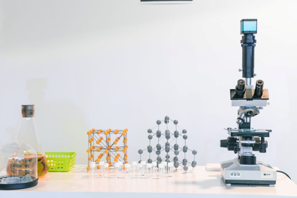 Uz galda izvietots laboratorijas aprīkojums: mikroskops ar kameru augšpusē, vairākas mazas paraugu pudelītes, divi ķīmiskie molekulu modeļi no savienotām lodītēm, stikla kolba ar korķi un zemā traukā ieliktas laboratorijas ierīces. Fons ir gaišs un minimālistisks