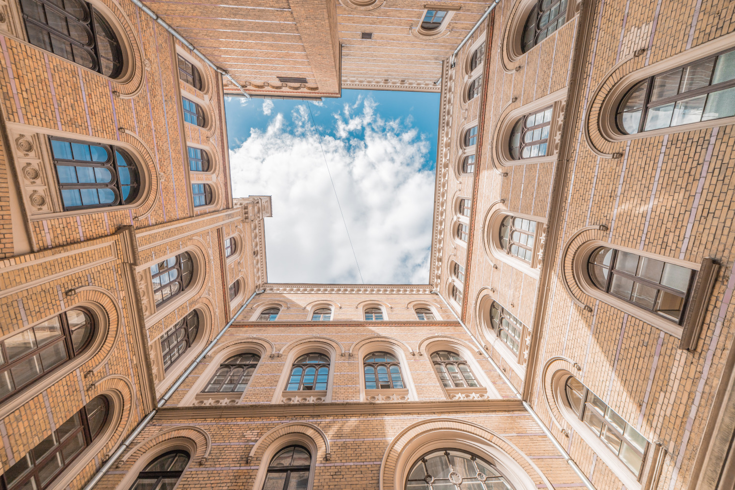 Skats uz augšu ēkas iekšpagalmā ar dzeltenīgu ķieģeļu fasādēm un arkveida logiem; četras sienas veido taisnstūrveida atvērumu, kura centrā redzamas debesis ar mākoņiem.