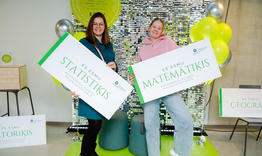 Divi cilvēki pozē pie dekoratīvas foto siena ar baloniem, katrs turot lielu kartona zīmi ar uzrakstiem “Es esmu statistiķis” un “Es esmu matemātiķis”. Fonā redzami arī citi līdzīgi studiju programmu plakāti un pasākuma atribūti.