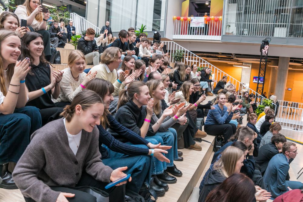 Plaša auditorija sēž Zinātņu mājas ātrijā , aktīvi aplaudējot un sekojot norisēm uz skatuves vai ekrāna.