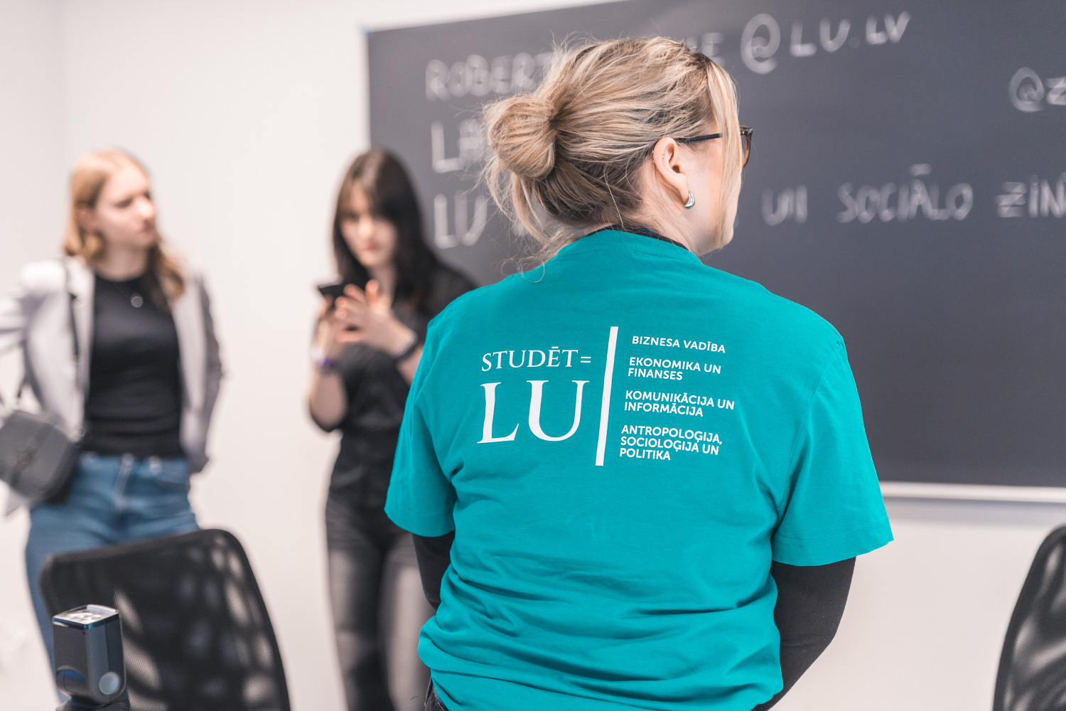Persona ar zaļu “Studēt LU” T–kreklu stāv auditorijā pie tāfeles, kur redzami uzraksti ar studiju informāciju, bet fonā atrodas vēl citas personas, kas iesaistās nodarbībā vai fotografē.