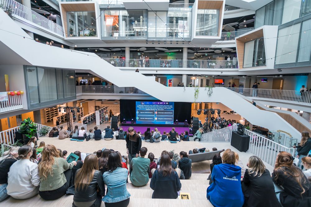 Plaša auditorija sēž daudzstāvu atvērta tipa ēkas centrālajā hallē, skatoties prezentāciju uz liela ekrāna; apkārt redzamas terases, galerijas un mūsdienīgs interjers ar stikla un metāla konstrukcijām.