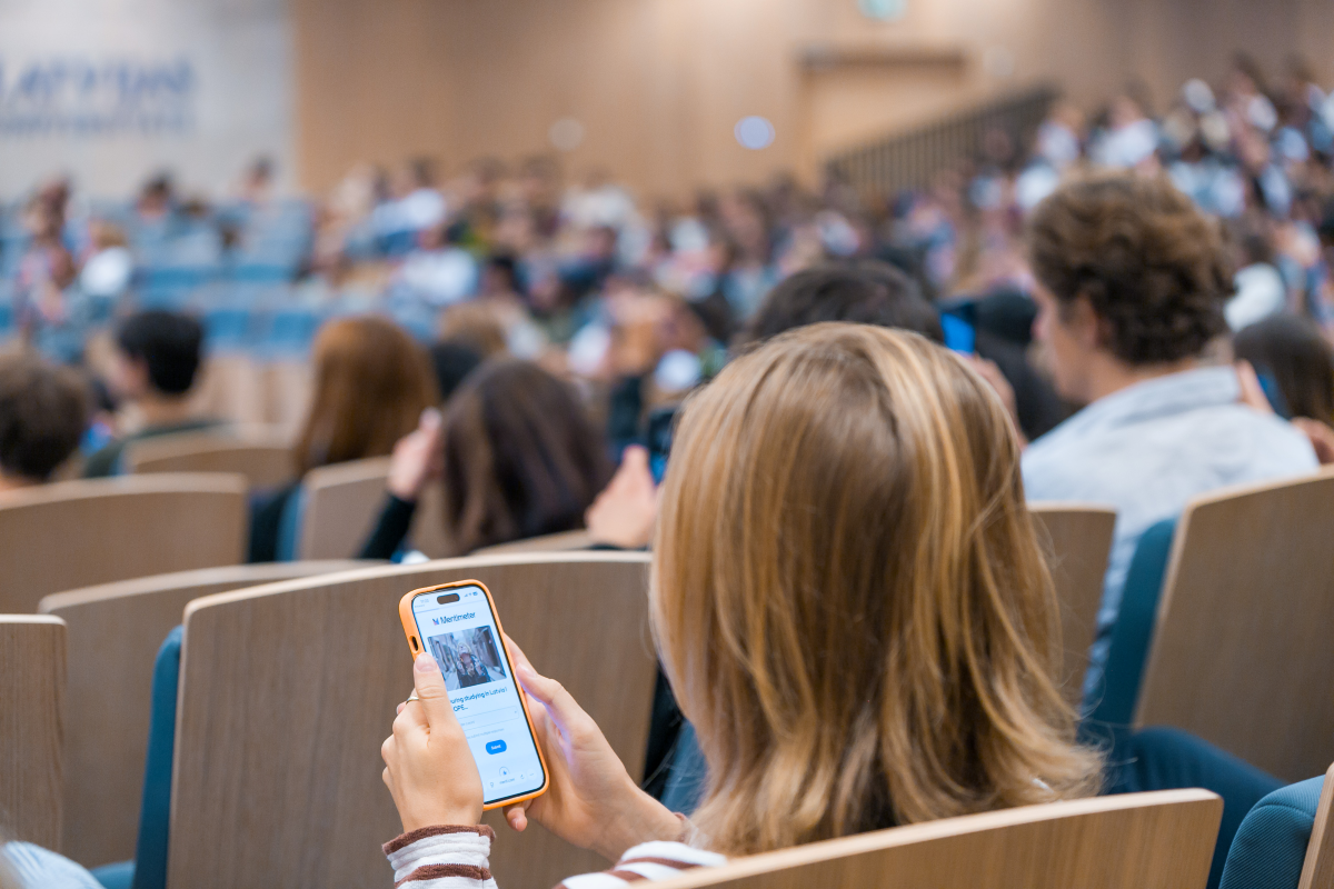 Alt teksts:  Students sēž auditorijā lekcijas vai pasākuma laikā. Priekšplānā redzams cilvēks, kurš sēdvietā izmanto viedtālruni, bet fonā redzama auditorija ar daudz apmeklētājiem un prezentācijas zona.
