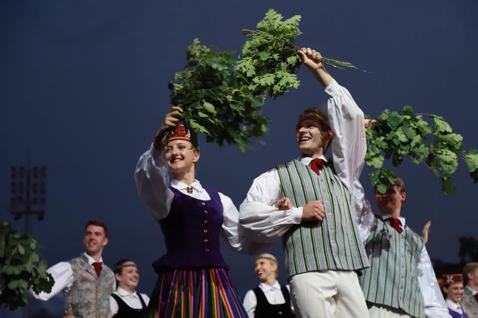 Tautas deju kolektīva dalībnieki tradicionālajos latviešu tautastērpos uzstājas, rokās turot ozolu zarus